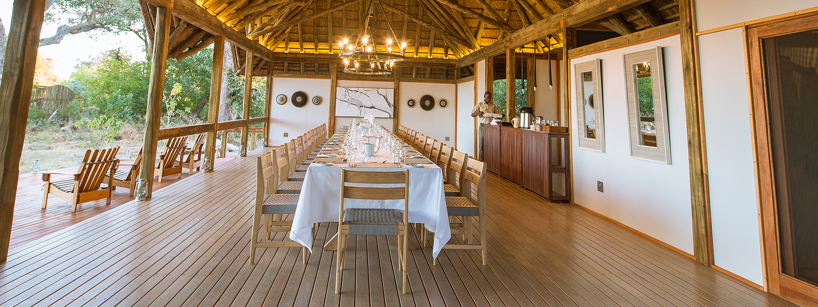 Long dining table set in an open-air wooden deck under thatched roofing.