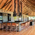 Interior view of Kanana Camp’s main lounge with modern bar, thatched roof, and décor inspired by the Okavango Delta, combining safari elegance with contemporary style.