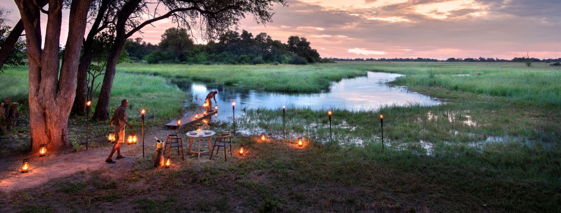 Romantic outdoor dinner setup beside a tranquil river in the savannah, illuminated by lanterns at sunset.
