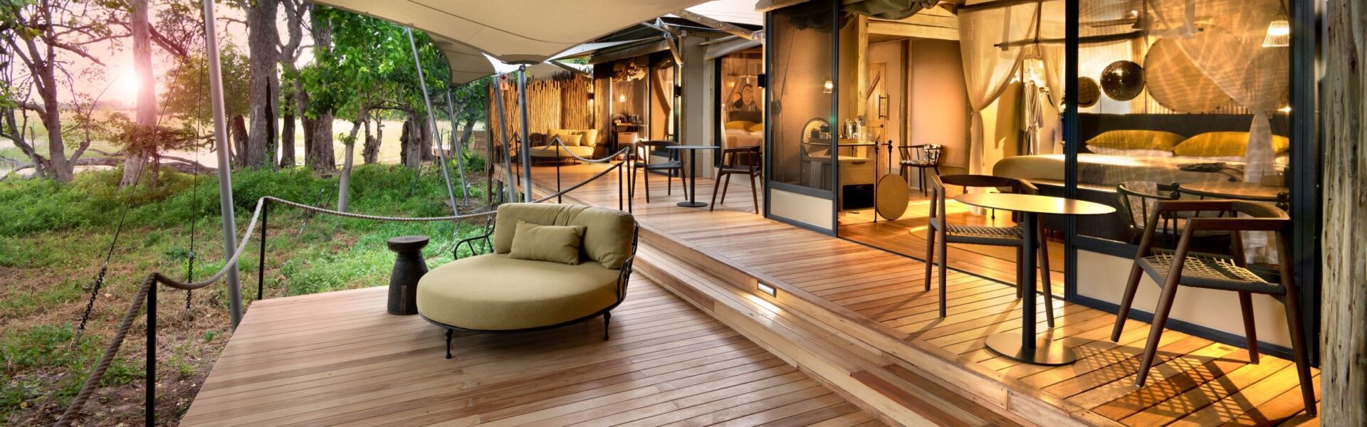 Spacious luxury tented suite with glass doors, elegant interiors, and a wooden deck surrounded by forest at sunset.