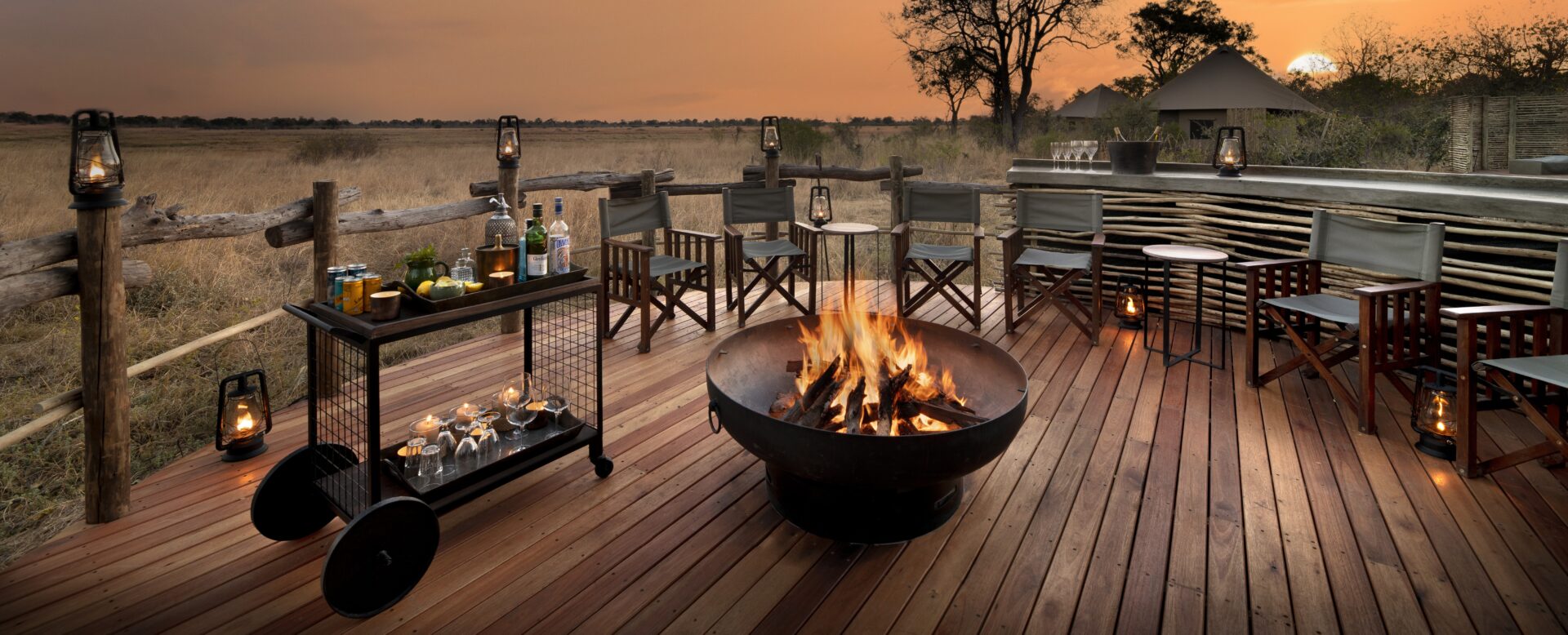 Outdoor firepit lounge area with director's chairs, a drinks trolley, and lanterns on a wooden deck overlooking the savannah at sunset.