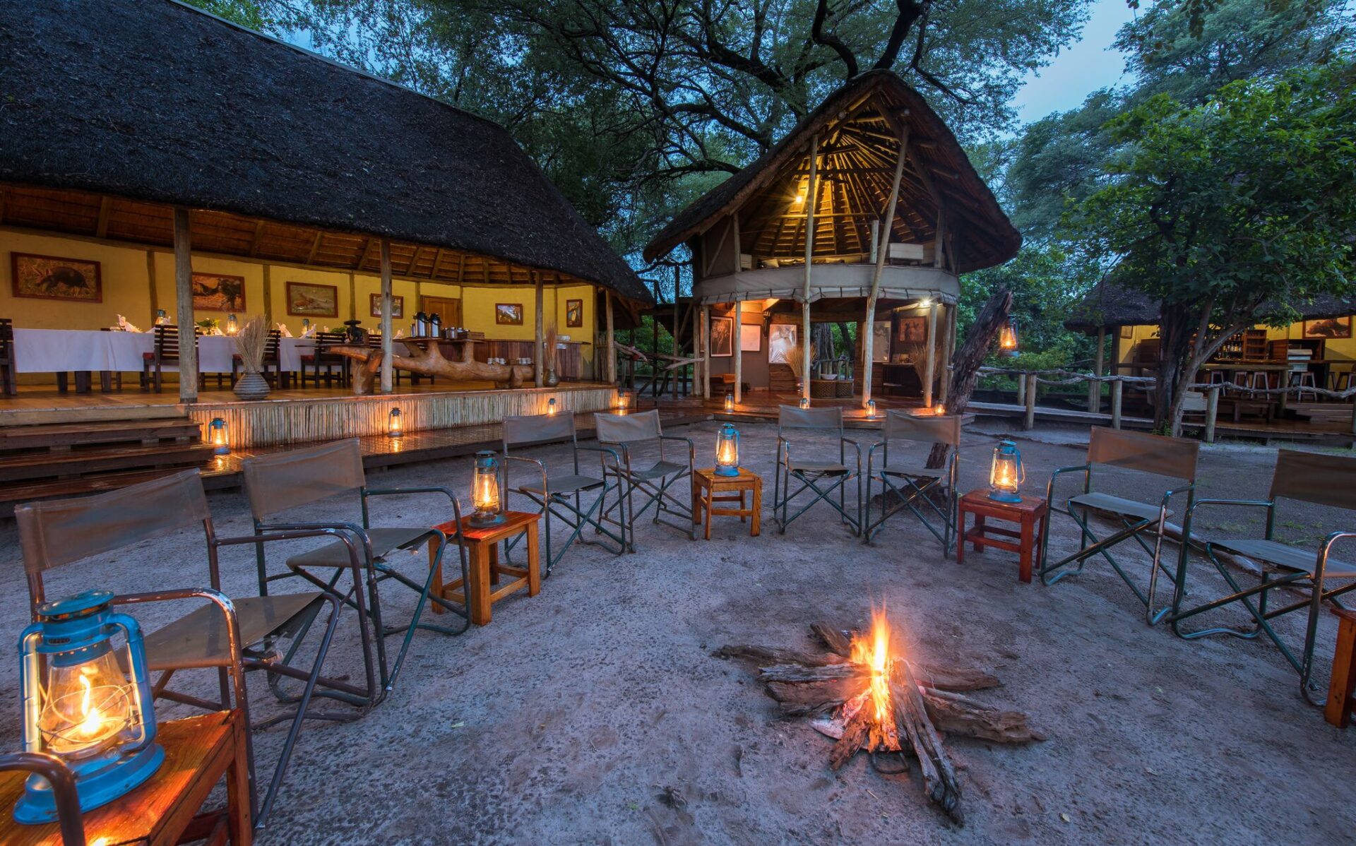 Campfire surrounded by chairs and lanterns outside thatched lodges at Lagoon Camp.