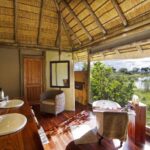 Bathroom with twin sinks, bathtub, and view of lagoon through thatched roof window.