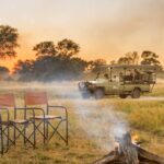 Safari vehicle parked near a campfire with chairs and sunset in the background.