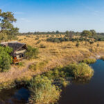 Aerial view of safari camp beside a lagoon with trees and golden grasslands.