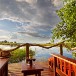 Two wooden chairs on a deck overlooking water with wine glasses on a table.