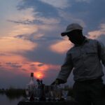 Two wooden chairs on a deck overlooking the river with two glasses of wine on a table.