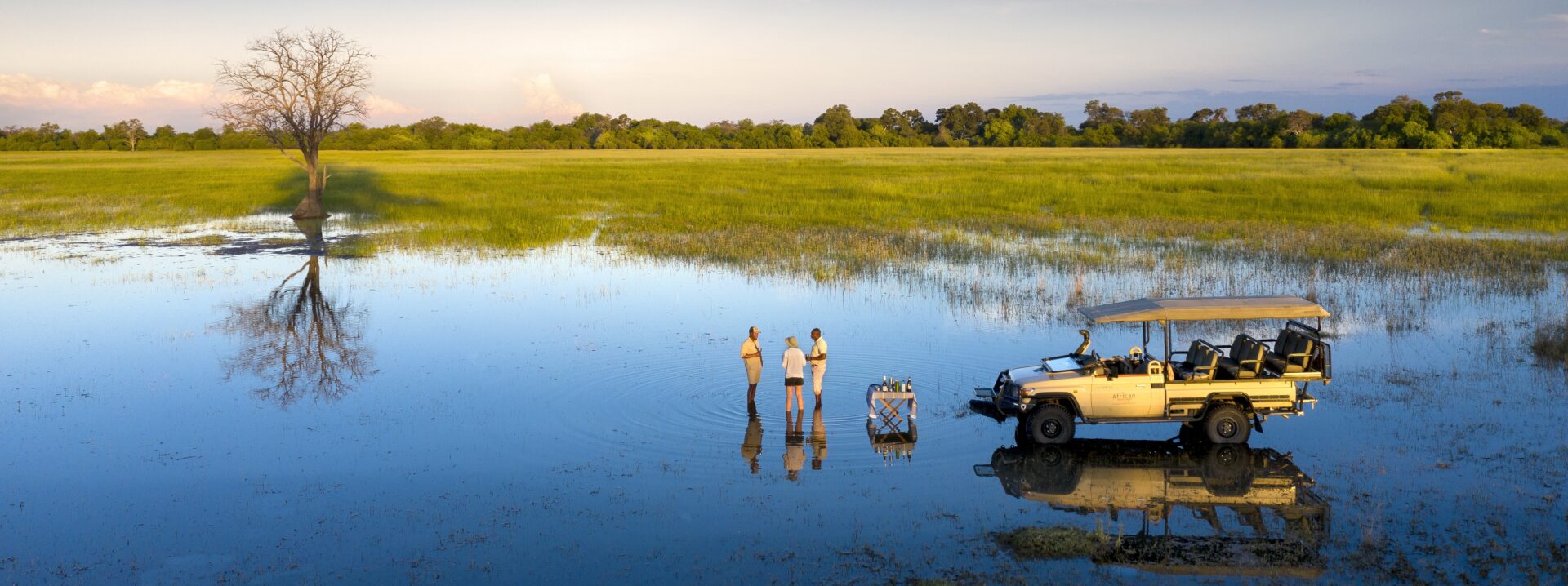 Three people stand in shallow water beside a safari vehicle enjoying drinks in the Okavango Delta at sunset.