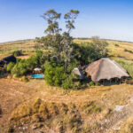 Aerial view of thatched safari lodges and swimming pool in grassy Botswana landscape.