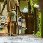 Person walking on a wooden path carrying several metal lanterns at an African safari camp.