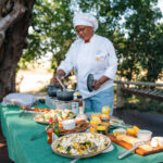 Chef cooking at an outdoor table set with various dishes under the trees.
