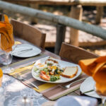 Outdoor dining table set with orange napkins, juice, and plated food.