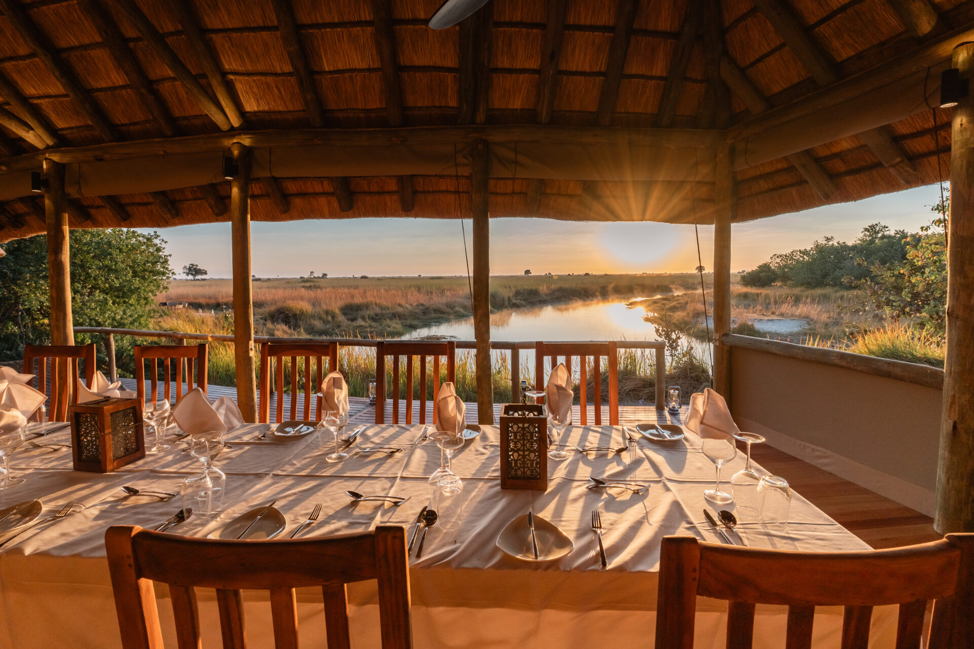 Dining table set under a thatched roof with sunset views over a river and grassy plains.