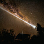 Person pointing a flashlight at the Milky Way in a star-filled night sky.