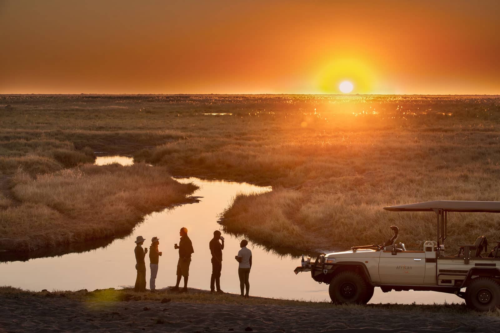 Linyanti Expeditions guests enjoying sunset drinks beside safari vehicle in Botswana.