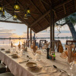 Lantern-lit dining table under thatch roof beside the lagoon at Pom Pom Camp.