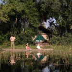 Mokoro with guide and guests passing in front of Pom Pom Camp tent on the lagoon.