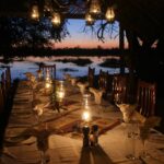 Outdoor dinner table lit by lanterns beside the water at Pom Pom Camp.
