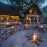 Campfire surrounded by chairs and lanterns outside thatched lodges at Lagoon Camp.