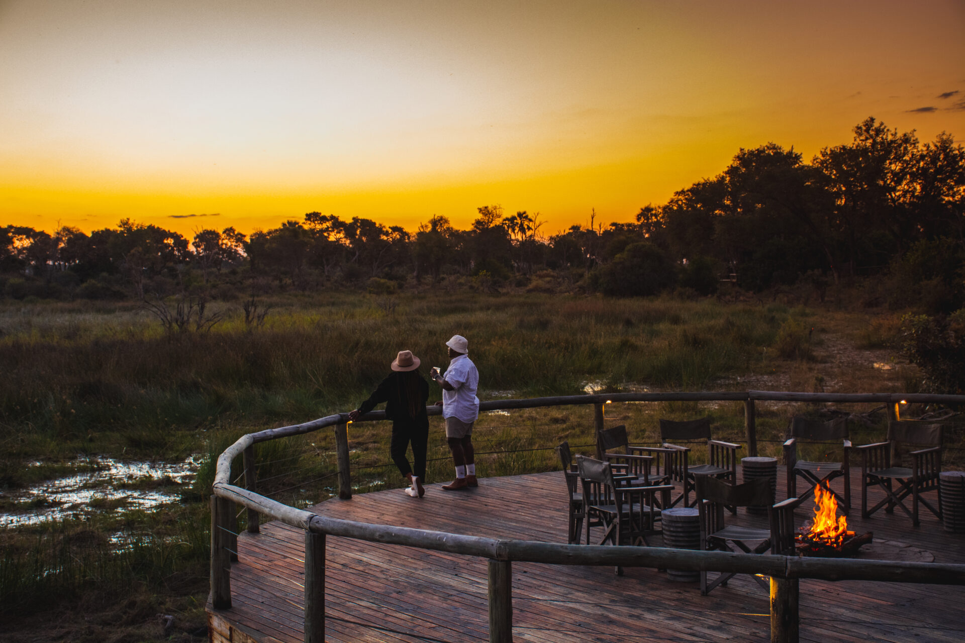 Two guests standing on the wooden deck with campfire seating overlooking the Delta at sunset at Mma Dinare Camp.