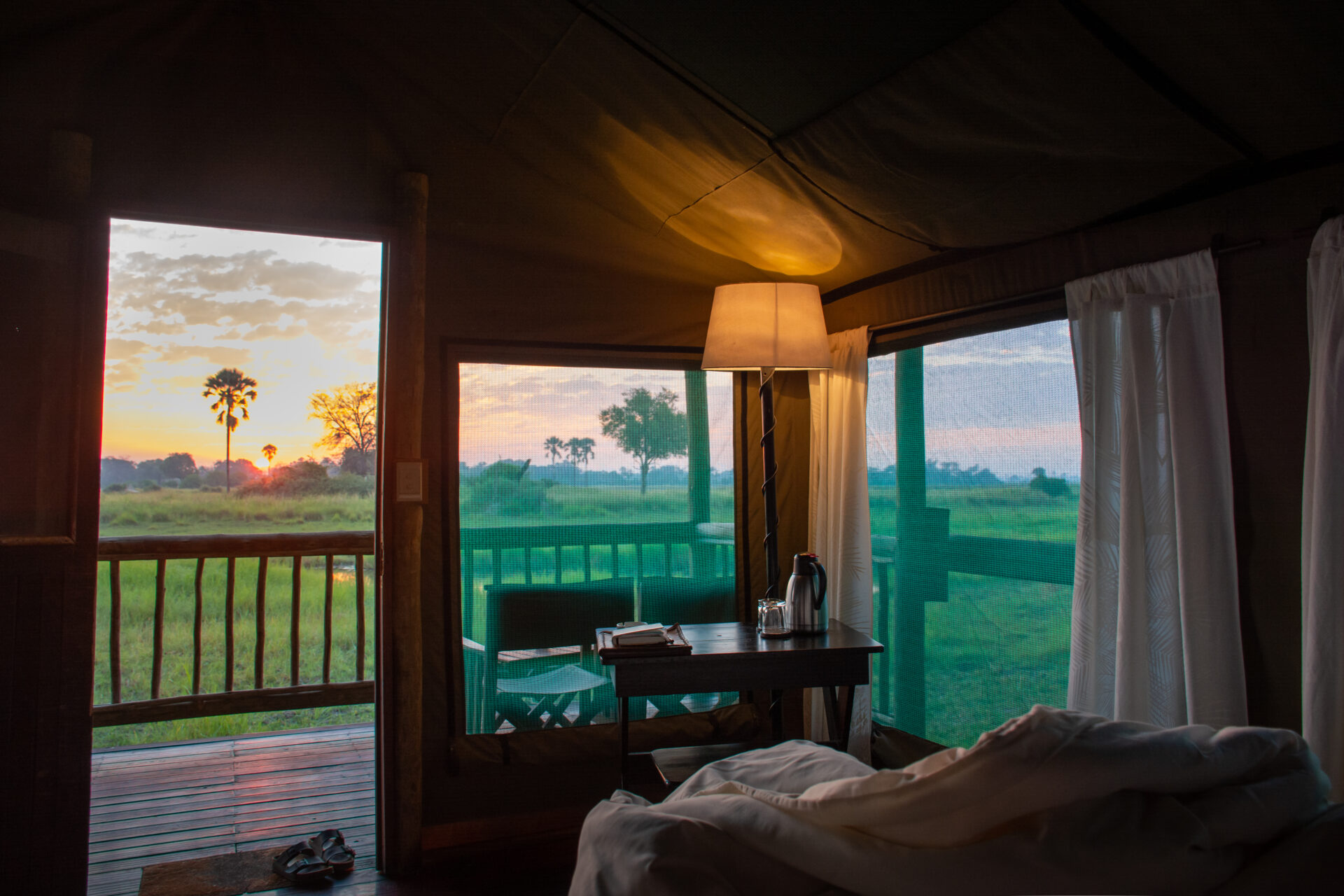Interior of a tented safari room with open curtains showing sunrise over grassy plains at Moremi Crossing.