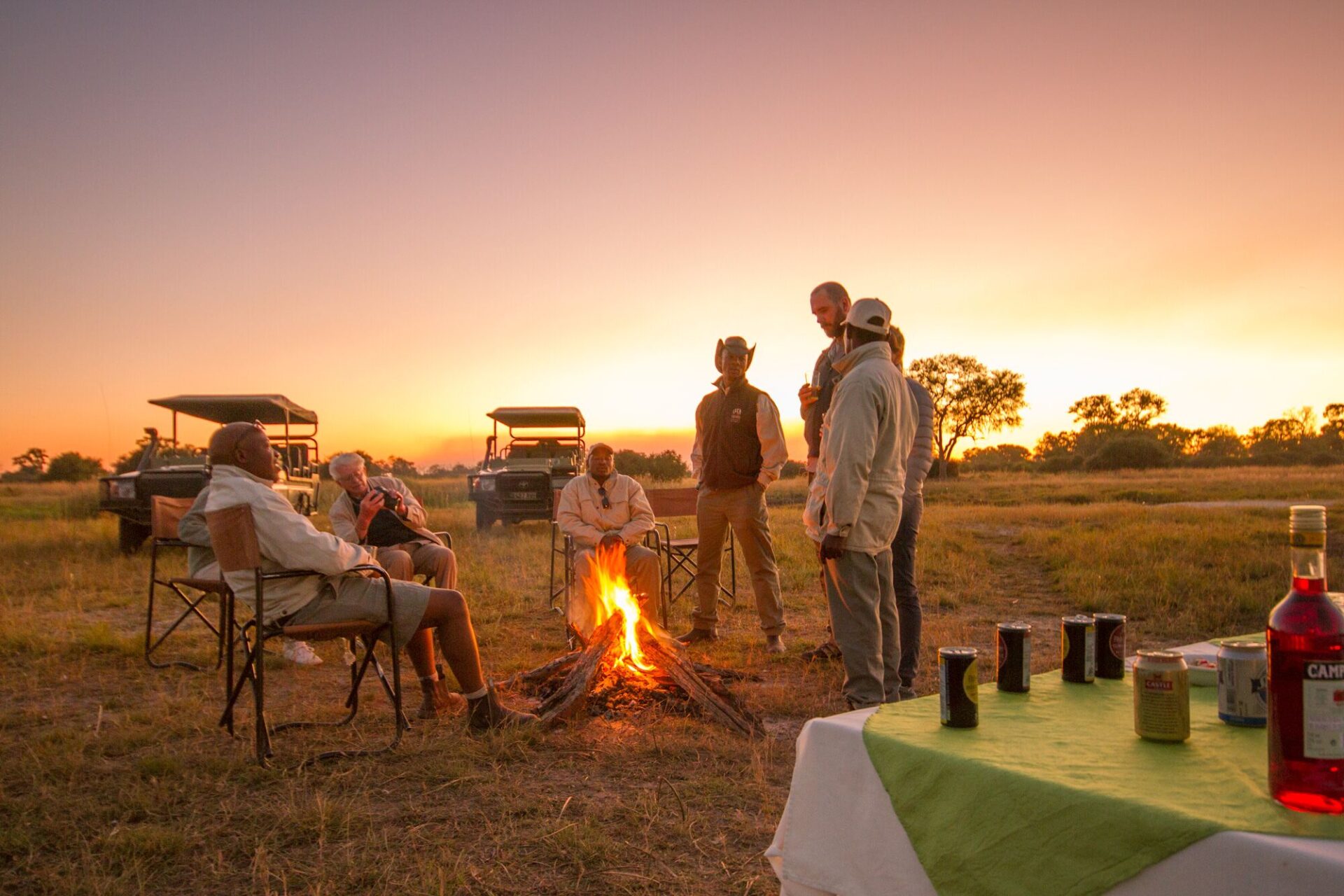 Group of safari guests sitting and standing around a campfire at sunset with drinks set on a nearby table at Lebala Camp.