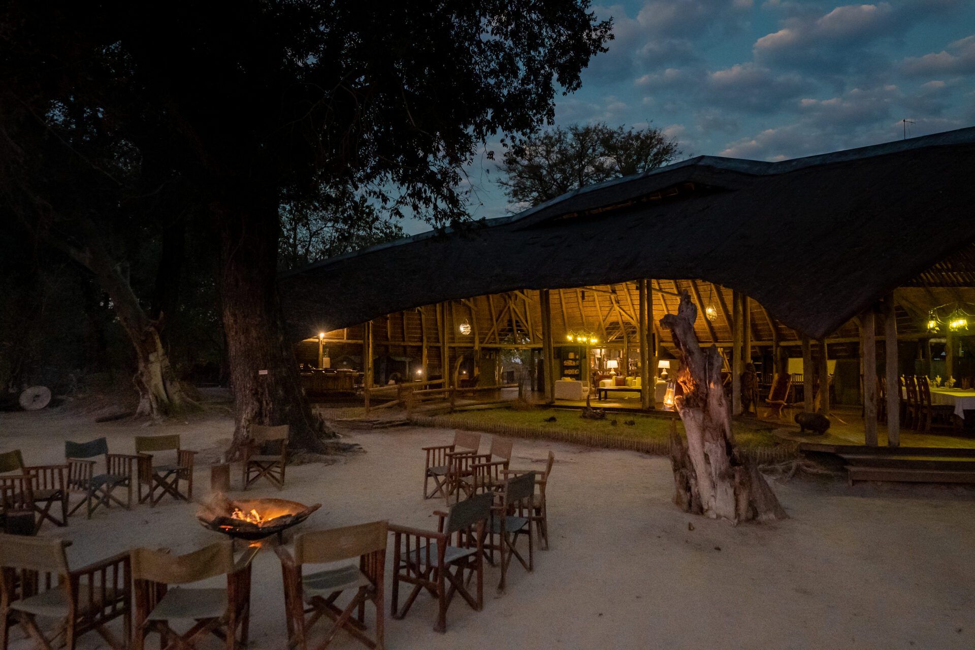 Outdoor seating area with wooden chairs around a firepit and a warmly lit thatched lounge at Pom Pom Camp in the evening.