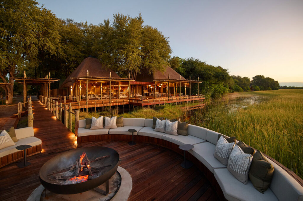 Curved fire deck seating at Sediba Sa Rona overlooking the Okavango Delta at dawn, with a glowing fire pit, golden grasslands and soft morning light.