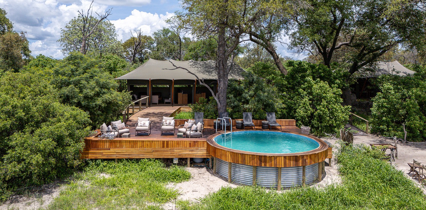 A luxury safari lodge with a raised wooden deck, comfortable lounge chairs, and a circular swimming pool surrounded by lush green trees and vegetation.