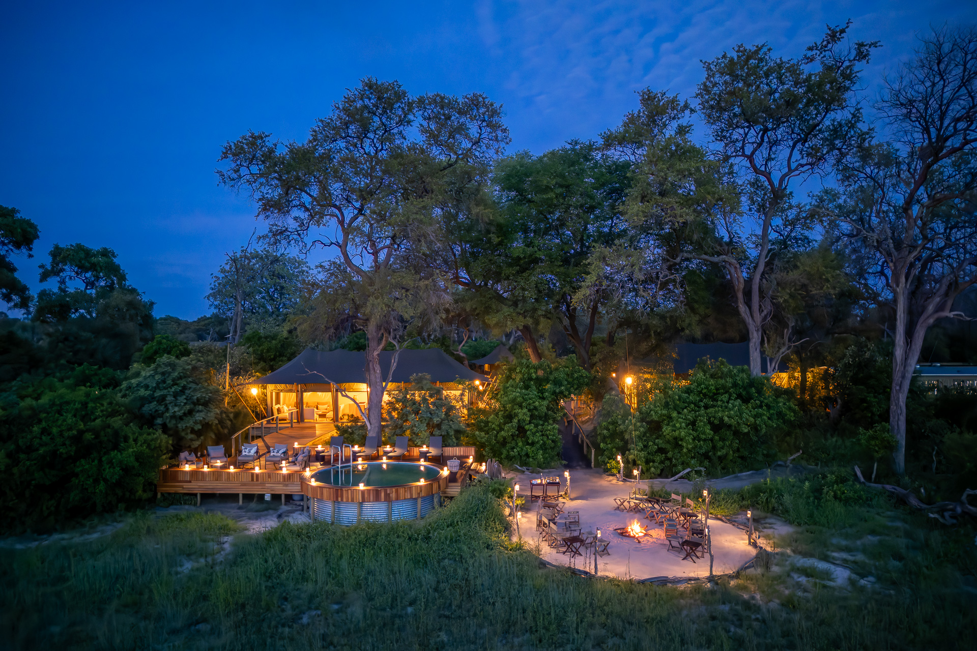 Luxury safari lodge lit up at dusk with a firepit and seating area surrounded by trees.