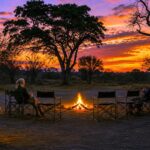 Guests seated around a campfire at sunset in a safari camp surrounded by trees.